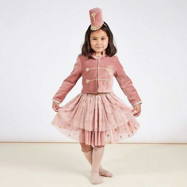 A young girl wears a Meri Meri Pink Soldier Costume with gold lamé details and a starry skirt, complimented by a small crown on her head, posing with a smile against a plain background.