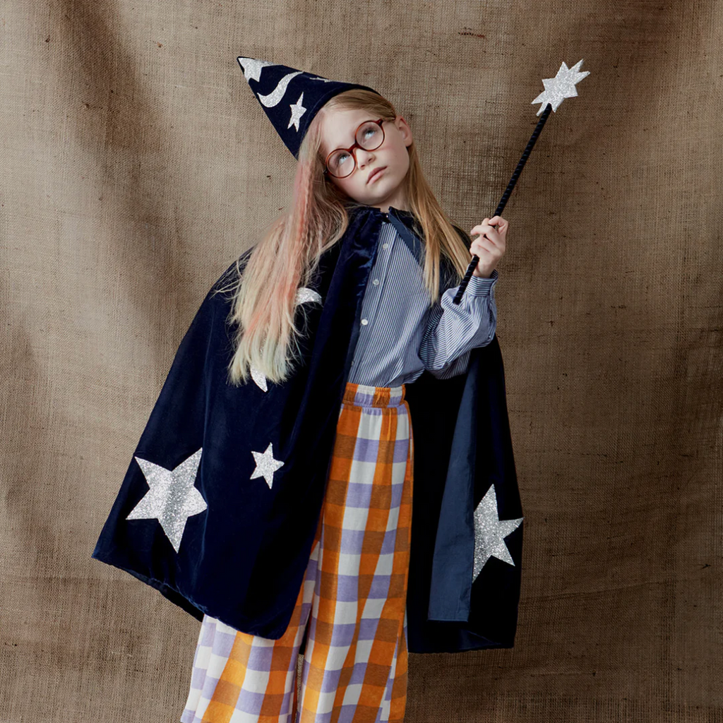 A young girl dressed in the Meri Meri Blue Velvet Wizard Costume, with a starry hat and a blue velvet cape, holding a wand, poses against a textured brown backdrop.