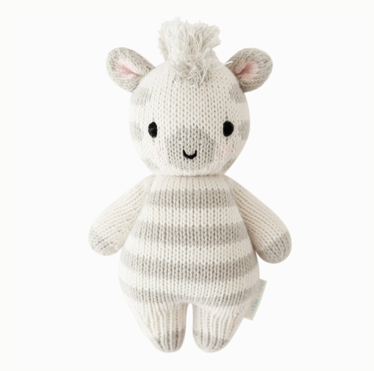 A Cuddle + Kind Baby Zebra, hand-knit from Peruvian cotton yarn, featuring grey stripes on a white body, styled with a fluffy tuft on top of its head and embroidered facial features, standing
