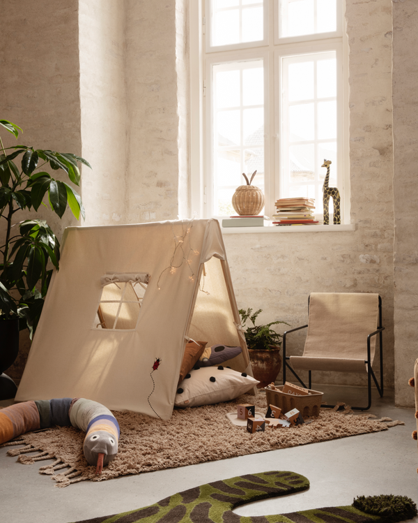 A cozy Ferm Living Tent - Ladybird Embroidery - Natural in a sunlit room with a beige rug, scattered toys, and a comfortable chair by the window. Organic cotton cushions inside the tent add to the inviting ambiance.