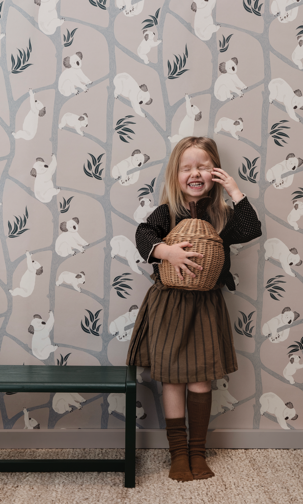 A young girl laughs while holding a Braided Apple Storage, standing against a wallpaper background featuring a pattern of playful polar bears. She wears a brown dress and long socks.