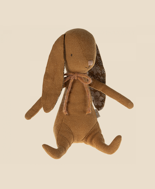 A Maileg Bunny - Ocher with long, floppy ears and a small embroidered nose. The bunny wears a brown ribbon tied around its neck and has patterned fabric on the insides of its ears. Made from recycled polyester, it has magnets in its hands, perfect for cuddles. The toy is seated against a plain beige background.