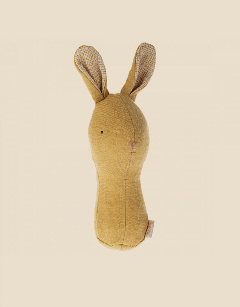 A Maileg Lullaby Friend Rattles, Bunny - Dusty Yellow with beige fabric and long ears lined with a checkered pattern. The toy has minimalistic embroidered facial features, perfect for little ones, and includes a baby rattle inside. Packaged in a charming gift box, it makes an ideal present. Background is a plain off-white color.