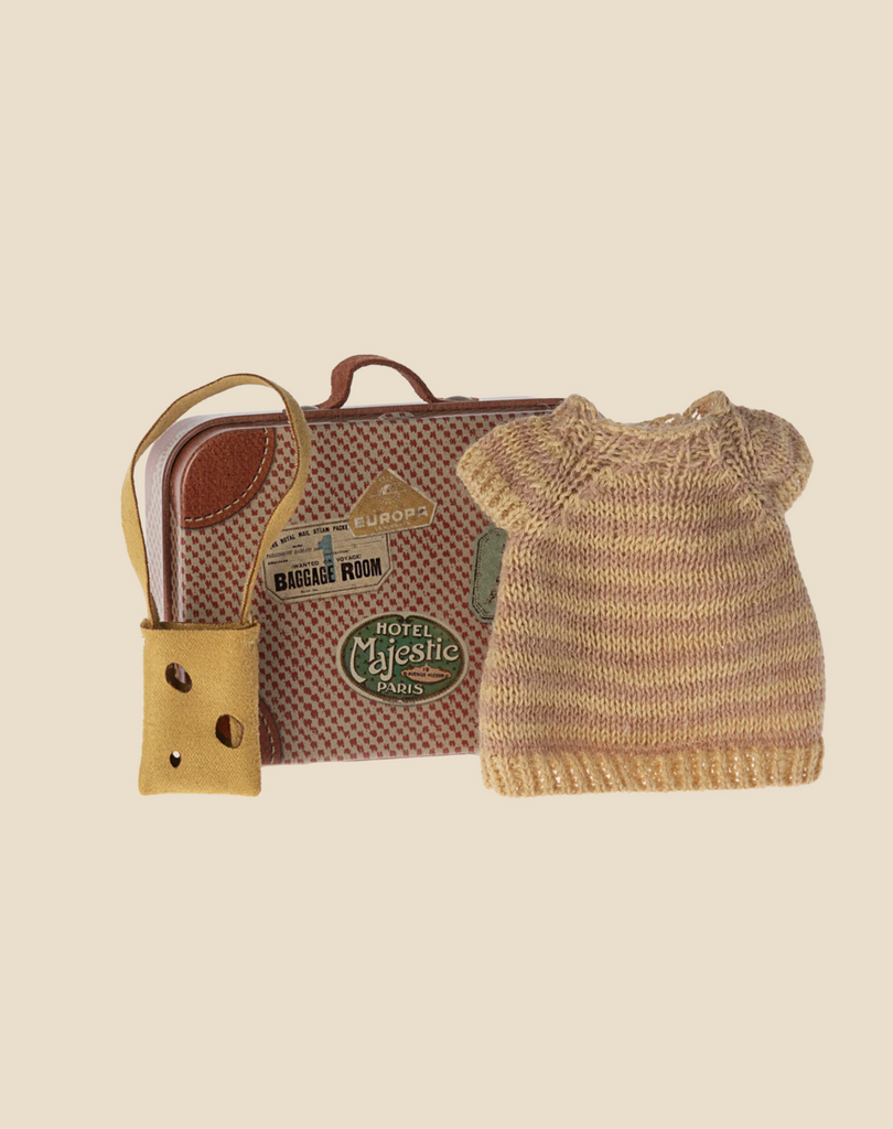 A small knitted sweater and a miniature suitcase with travel stickers are placed next to a yellow luggage tag. The Maileg Knitted Dress And Bag In Suitcase, Big Sister Mouse, ideal for a big sister/brother toy, features stickers from various locations like "Hotel Majestic Paris" and "Baggage Room." The background is solid beige.