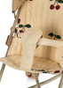 Close-up image of a Konges Sloejd Zoe Doll Stroller - Cherry with beige seat featuring cherry print fabric. The seat includes padded straps and a secure harness system. The frame, made of light-colored metal, incorporates double wheels for stability and a foldable shade for sun protection.