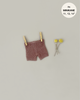 Minikane Doll Clothing | Vito Shorts in Marl Caramel Knit hang from two wooden clothespins against a light grey background. Beside the shorts, a small bouquet of three yellow pom-pom flowers adds a touch of charm. A round label in the top right corner reads "fits MINIKANE 11, 13, 14".