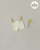 A pair of Minikane Doll Clothing | Vito Shorts in Cream Knit is clipped to a clothesline with two wooden clothespins. The background is plain beige. Two small yellow flowers are positioned on the right side of the shorts. Text in the top right reads, "fits MINIKANE 11, 13, 14” – perfect for Minikane Babies or Gordis dolls.