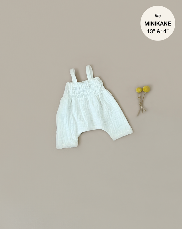 A Minikane Doll Clothing | Bonnie Jumpsuit in Cream for Minikane Gordis dolls measuring 13 to 14 inches is positioned against a beige background. The romper, made of double gauze, has shoulder straps and a gathered bodice. Beside it, two small yellow ball-shaped flowers tied together lie flat. The text indicates it fits "MINIKANE 13" & 14".