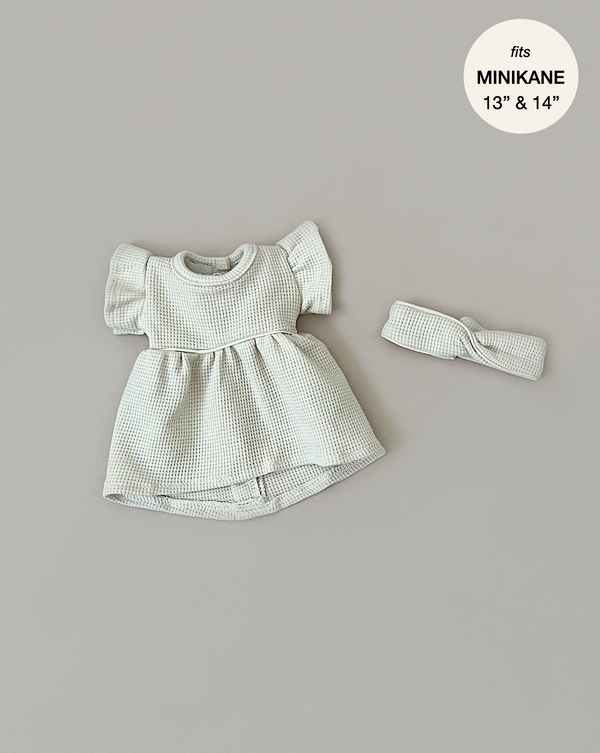 A small Minikane Doll Clothing | Linen Dress and Headband Set lies on a neutral background. The dress features flutter sleeves and a high-low hem, perfect for Minikane Gordis dolls. A circular label at the top right reads "fits MINIKANE 13” & 14”".