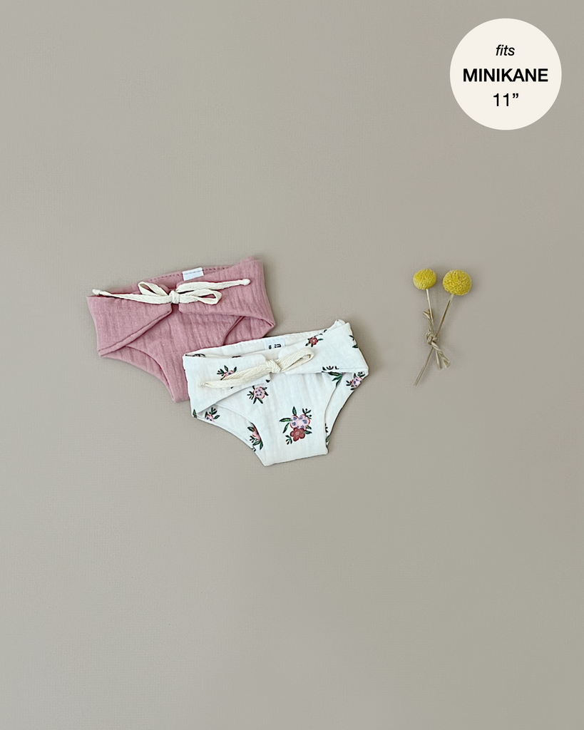 Two pairs of Minikane Doll Clothing | Set of 2 Diapers in Petal Pink and Floral lie against a neutral background. One pair is pink with a white tie, and the other is white with a floral pattern. A small bouquet with two yellow billy button flowers is placed to the right. Perfect for your Minikane Babies, these could also resemble double gauze diapers.