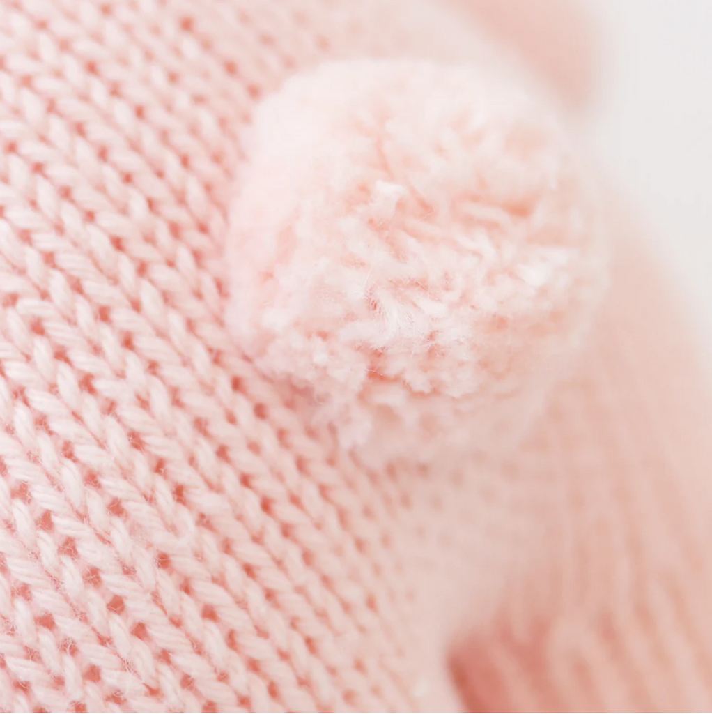 Close-up image of a soft, light pink knitted fabric made from heirloom-quality natural Peruvian cotton yarn, with a fluffy pom-pom attached. The texture of the knit and the details of the fuzzy material are clearly visible, emphasizing the cozy and delicate appearance of the Cuddle + Kind Baby Rabbit, Powder Pink.