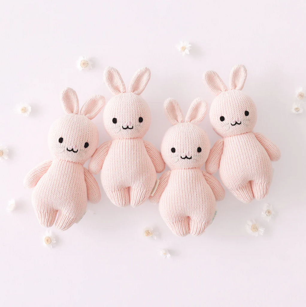 Four identical Cuddle + Kind Baby Rabbit, Powder Pink dolls, crafted from natural Peruvian cotton yarn, stand in a row on a pale pink background. With smiling faces, black eyes, and long ears, these heirloom-quality hand-knit baby animals are surrounded by small white flowers that add a delicate decorative touch.