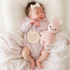 A baby sleeps on a soft, patterned blanket. The baby wears a light pink knit outfit made from heirloom-quality Peruvian cotton yarn and a cream-colored bow headband. In one arm, the baby holds a Cuddle + Kind Baby Rabbit, Powder Pink toy. A wooden sign next to their chest reads "HELLO WORLD.