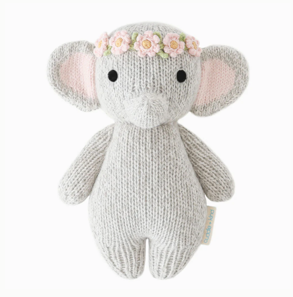 A Cuddle + Kind Baby Elephant, Blush Floral with large ears and black eyes. The elephant is wearing a pink flower crown on its head. Made from Peruvian cotton yarn, it has a hand-knit texture and a tag on its side, ensuring heirloom-quality charm.