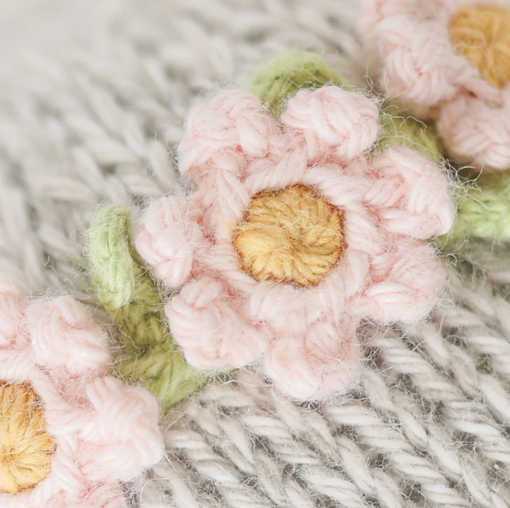 Close-up of a knitted pattern featuring delicate pink flowers with yellow centers and green leaves, set against a textured beige background. The knitting, crafted with Cuddle + Kind Baby Elephant, Blush Floral yarn, showcases fine details and heirloom-quality craftsmanship.