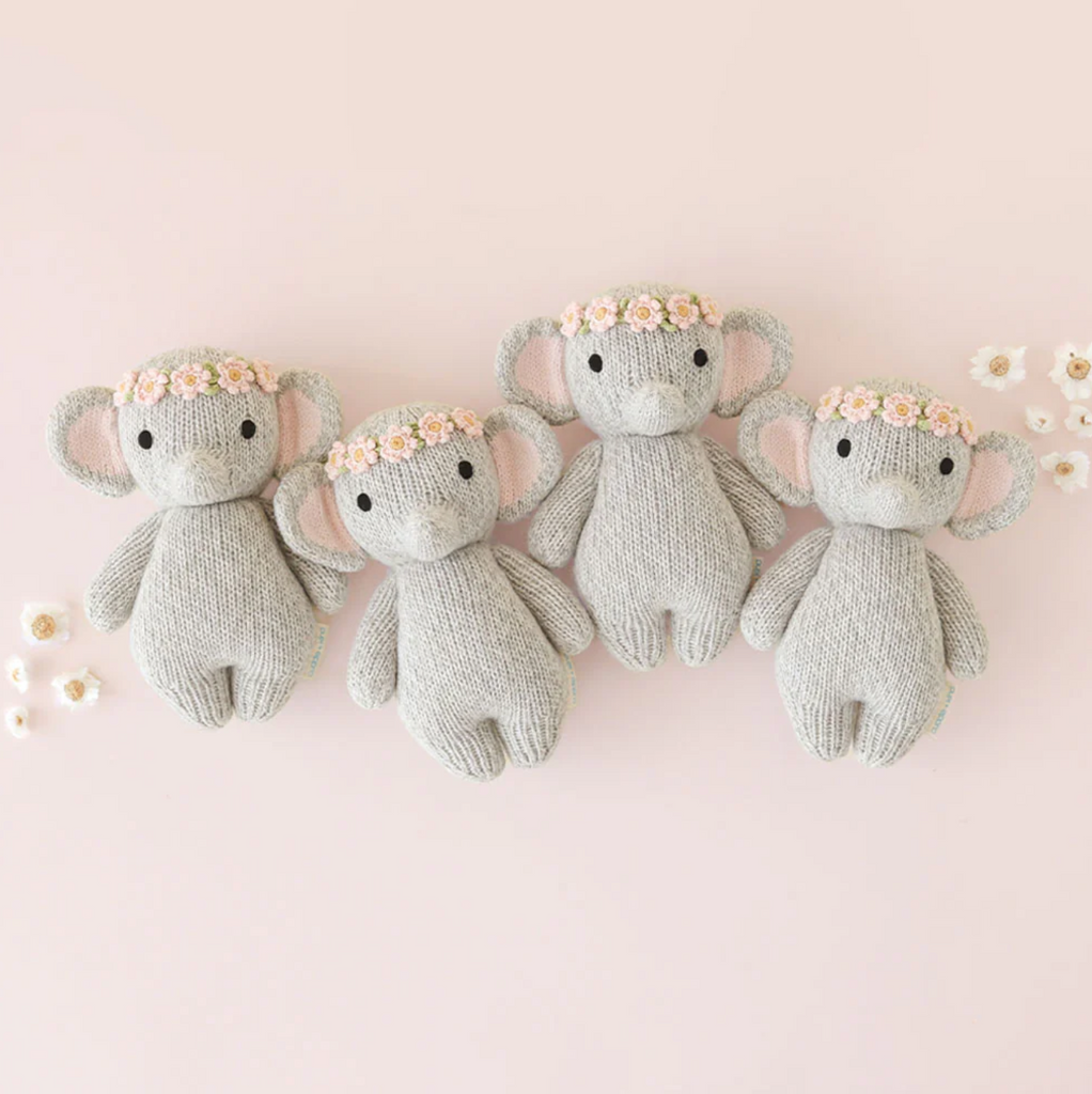 Four identical Cuddle + Kind Baby Elephant, Blush Floral plush toys, each adorned with a pink floral crown, are neatly lined up on a pale pink background. Small white flowers are scattered around them, enhancing the charming and whimsical scene crafted from heirloom-quality Peruvian cotton yarn.