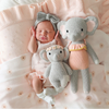 A newborn baby wearing a pink outfit and a grey headband lies on a pink blanket. The baby is nestled between two heirloom-quality, hand-knit baby animals made from soft Peruvian cotton yarn. One small and one large, the Cuddle + Kind Baby Elephant, Blush Floral dolls in pink and grey attire create a cozy and serene scene.