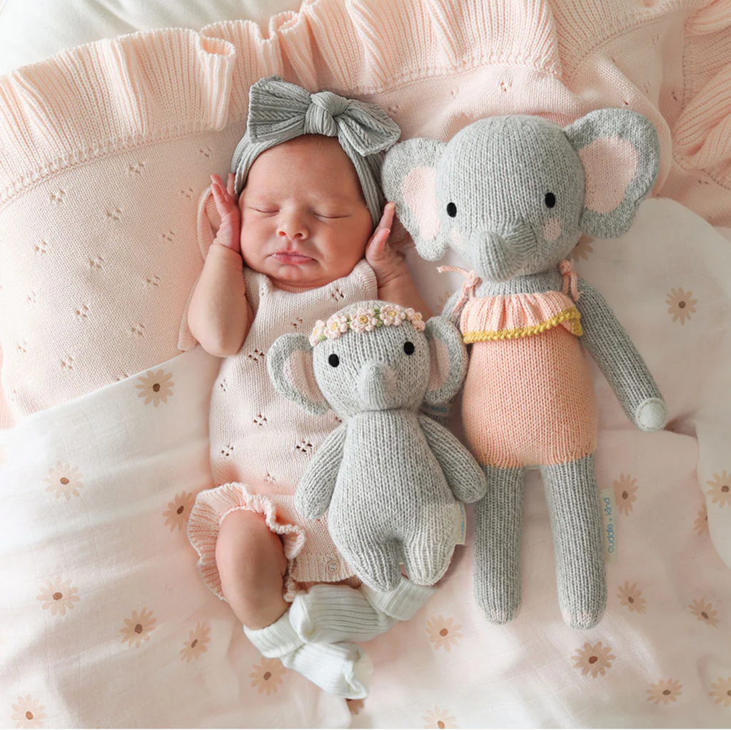 A newborn baby wearing a pink outfit and a grey headband lies on a pink blanket. The baby is nestled between two heirloom-quality, hand-knit baby animals made from soft Peruvian cotton yarn. One small and one large, the Cuddle + Kind Baby Elephant, Blush Floral dolls in pink and grey attire create a cozy and serene scene.