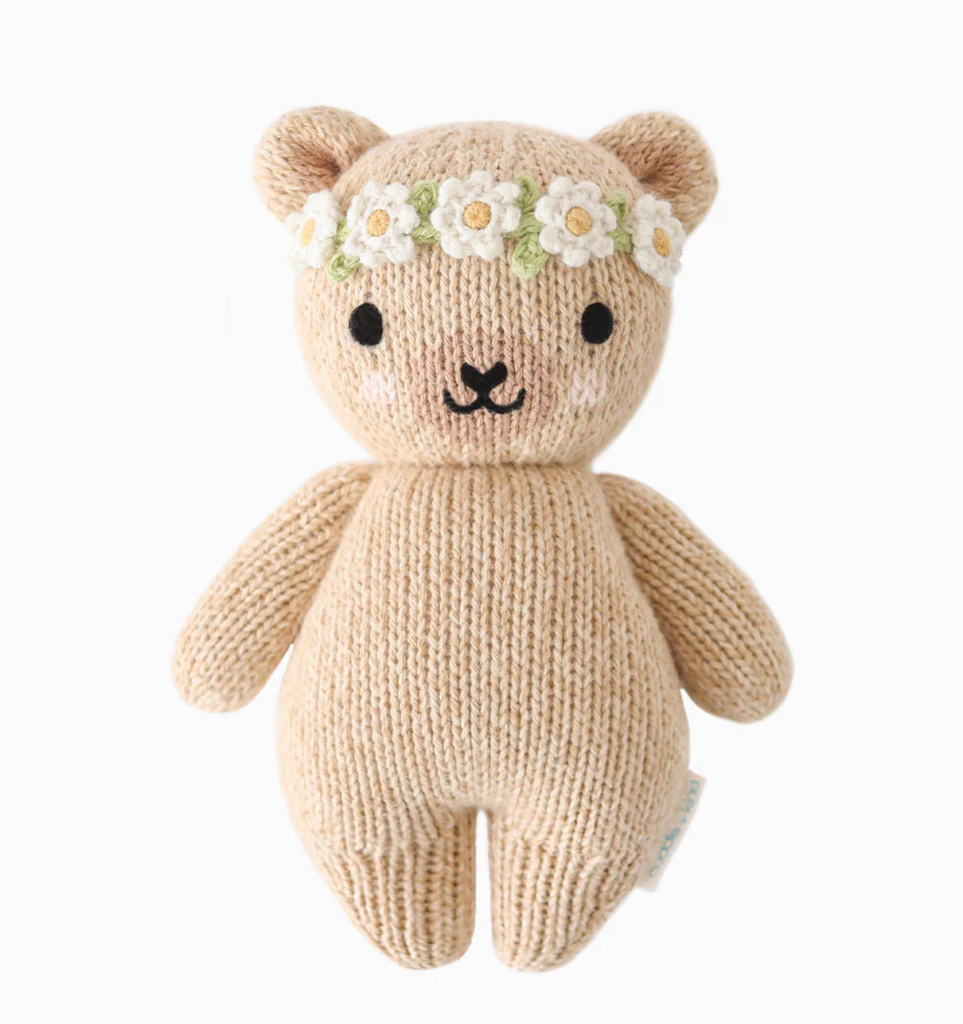 A Cuddle + Kind Baby Honey Bear, Ivory Floral made of natural Peruvian cotton yarn with a beige body, black eyes, and a small black nose and mouth. It is adorned with a crocheted flower crown of white daisies with yellow centers and green leaves. This heirloom-quality piece sits against a white background.