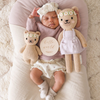 A sleeping baby rests on a soft, cushioned surface, dressed in a lavender long-sleeve top, white shorts, and a white headband with a bow. Flanked by two heirloom-quality Cuddle + Kind Baby Honey Bear, Ivory Floral crafted from natural Peruvian cotton yarn, a wooden sign reading "hello world" lies beside the baby.