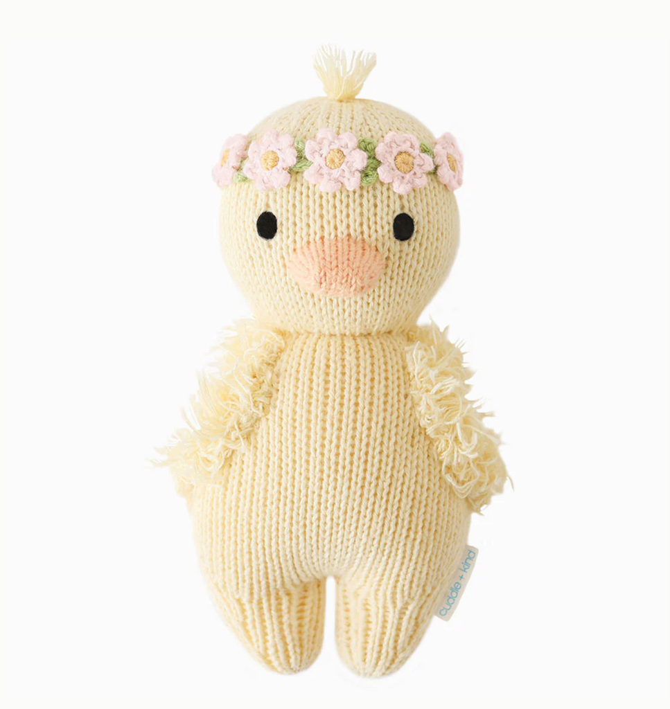 A Cuddle + Kind Baby Duckling, Blush Floral crafted from heirloom-quality Peruvian cotton yarn, featuring a light yellow body and small, black eyes. Adorned with a pink flower crown on its head and fluffy-textured wings, this charming toy also has a tag on its side.