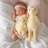 A newborn baby, wearing a white floral headband and light pink outfit, is peacefully sleeping on a light pink blanket. The baby is cuddling two Cuddle + Kind Baby Duckling, Blush Floral plush toys of different sizes, both adorned with flower crowns. Made from heirloom-quality Peruvian cotton yarn, the scene is soft and cozy.