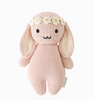 A pink handcrafted Cuddle + Kind Baby Bunny, Rose With Ivory Floral with long ears, black eyes, and a small smiling mouth. The bunny is wearing a white and green flower crown around its head. It has a soft, textured appearance, and a label is visible on its side, making it a delightful collectible for enthusiasts.