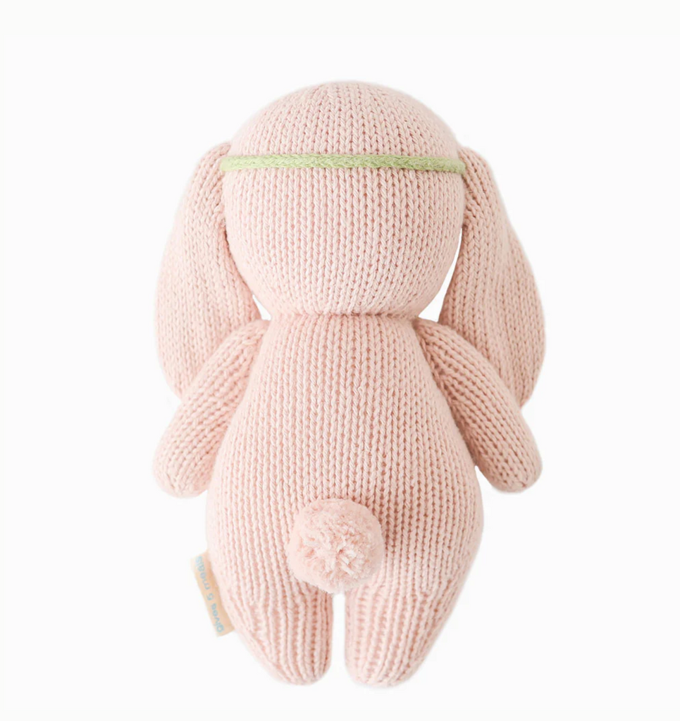 A soft, pink Cuddle + Kind Baby Bunny, Rose With Ivory Floral is shown from the back. Hand-knit from heirloom-quality Peruvian cotton yarn, the bunny has long floppy ears, a short, round tail, and a narrow green headband above its ears. The knitted texture is visible, and the toy has an overall cute and cuddly appearance.