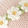 Close-up view of a handcrafted knitted fabric in soft pink with a decorative band featuring white crocheted flowers with yellow centers and green leaves. The intricate details of the knitted stitches and the delicate design of the flowers are clearly visible, making the Cuddle + Kind Baby Bunny, Rose With Ivory Floral truly collectible.