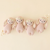 Four identical Cuddle + Kind Baby Bunny, Rose With Ivory Floral are lined up in a row against a light beige background. Each handcrafted bunny has floppy ears, embroidered facial features, and wears a floral headband made of little white daisies. These collectible baby animals are surrounded by scattered small daisies.