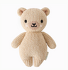 A Cuddle + Kind Baby Honey Bear made from natural Peruvian cotton yarn, featuring black eyes, a small black nose, and a smiling mouth. The collectible bear has round ears and is standing upright on a white background, embodying the charm of hand-knit baby animals.