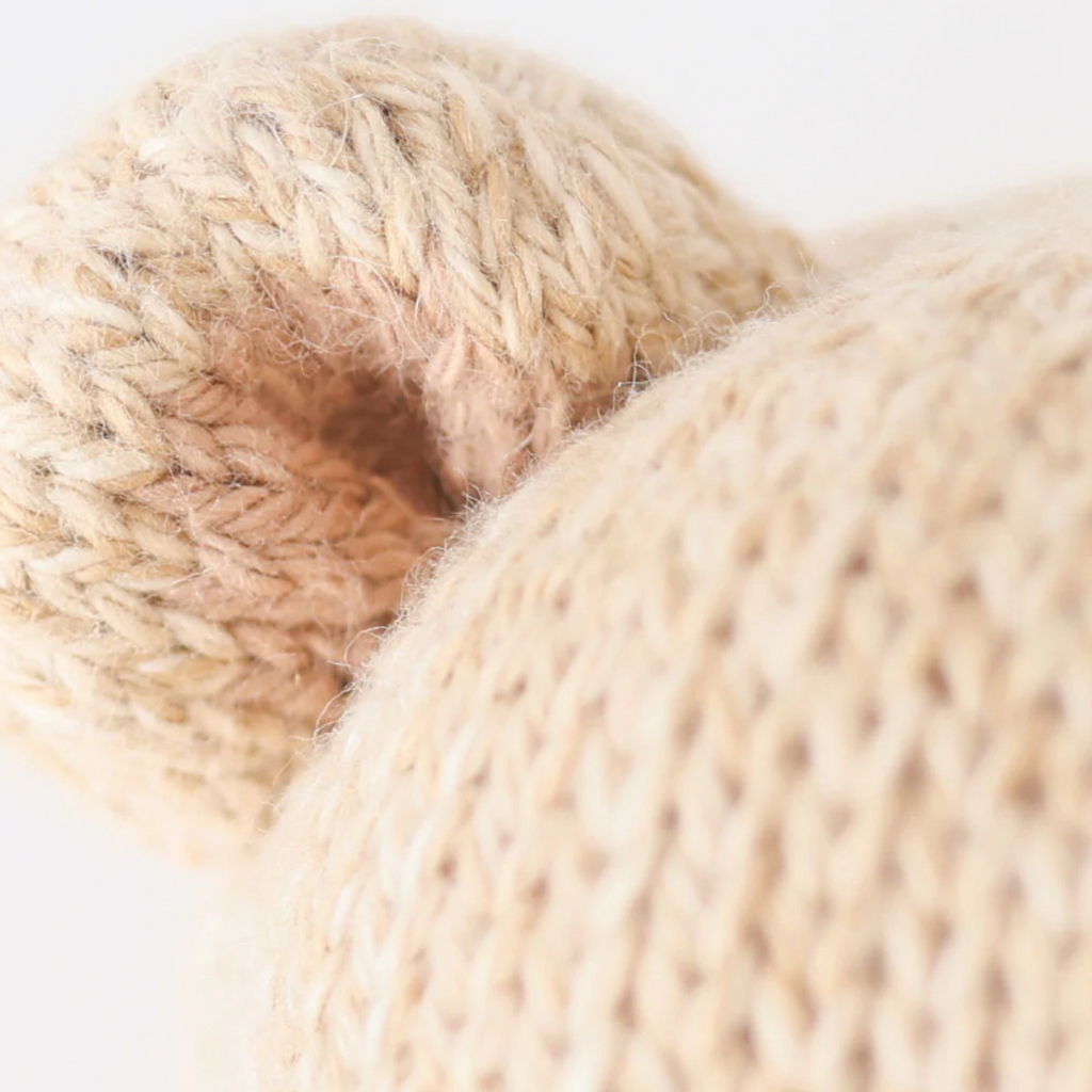 Close-up image of a knitted object with beige yarn, focusing on a rounded section resembling an ear. The texture shows detailed knit patterns, with some areas shaded slightly darker than others. Crafted from natural Peruvian cotton yarn, this Cuddle + Kind Baby Honey Bear showcases artisanal skill.