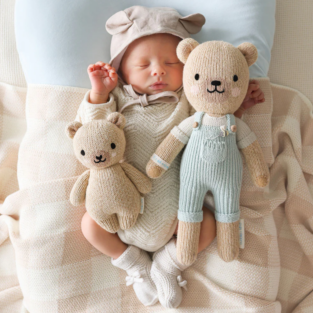 A sleeping baby, dressed in a beige knit outfit and hat with little ears, cuddles with two hand-knit baby animals. One Cuddle + Kind Baby Honey Bear is small and plain beige, made from natural Peruvian cotton yarn, while the other is larger and wears blue overalls. The baby and teddy bears are lying on a cream-colored blanket.