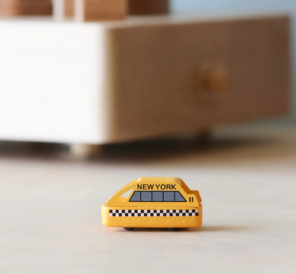 A Replacement Taxi For New York Music Box, featuring a small yellow cab with "New York" written on its side and a black-and-white checkered stripe, sits on a light-colored surface with a blurred wooden block structure in the background.