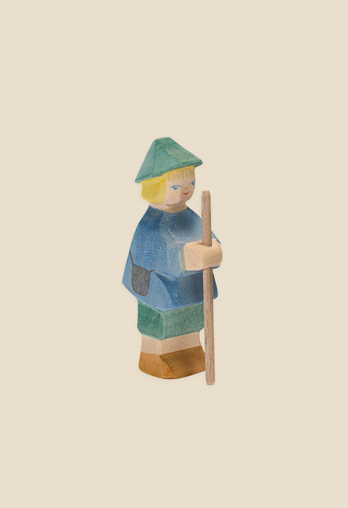 The Ostheimer Shepherd Boy is a handcrafted wooden figurine depicting a person with blonde hair, dressed in blue and green clothing, including a pointed hat, and holding a wooden staff. Set against a plain light beige background, this piece is perfect for imaginative play and embodies the exceptional quality of Ostheimer brand wooden toys.