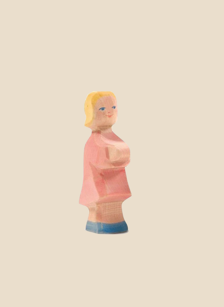 The Ostheimer Daughter in Pink Dress is a small wooden figure of a girl with short blonde hair, wearing a pink dress and blue shoes, holding a white object in front of her. Set against a plain, light brown background, this handcrafted toy from the Ostheimer brand encourages imaginative play in children.