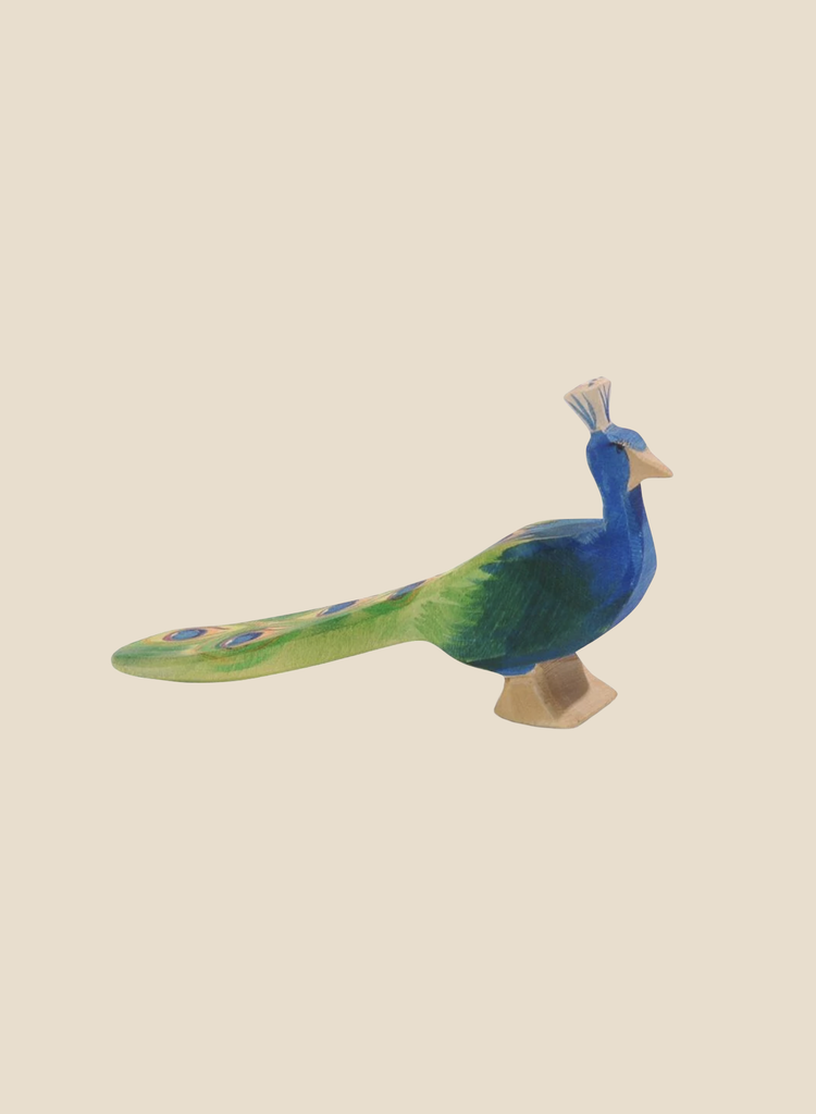 The Ostheimer Peacock, a colorful wooden bird figurine handcrafted from sustainably sourced materials, features a blue body, green tail, small yellow beak, and a decorative crest on its head. It stands against a plain beige background.