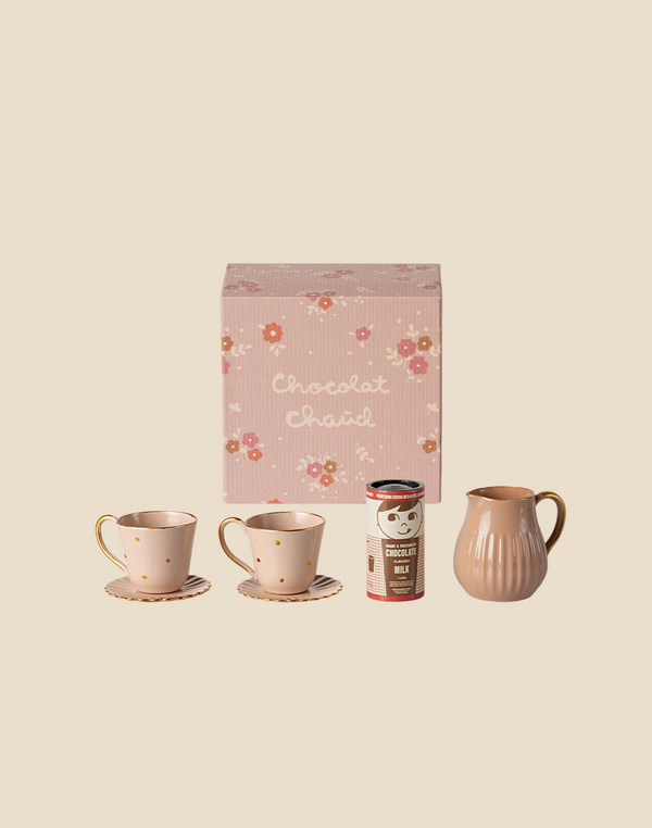 A pink floral box labeled "Chocolat Chaud" from the Maileg Hot Chocolate Set, Mini, includes two polka-dotted cups with saucers, a small pitcher, and a tin of hot chocolate milk powder. The background is a soft beige.