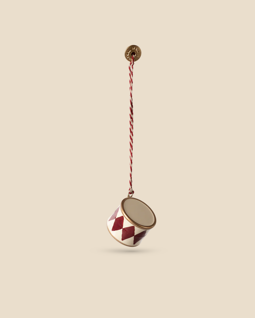 A Maileg Metal Ornament, Small Drum - Red features intricate handpainted details with red and white diamond patterns hanging from a twisted cord, set against a plain beige background.