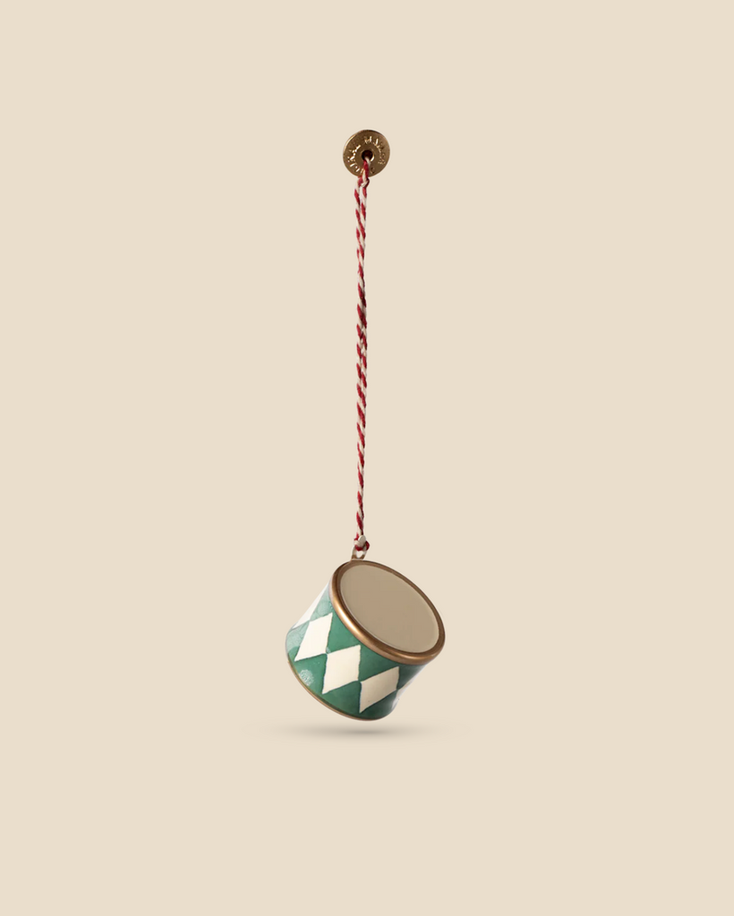 A Maileg Metal Ornament featuring a small dark green drum with handpainted details and a green and white diamond pattern hangs from a twisted red and white string against a beige background.