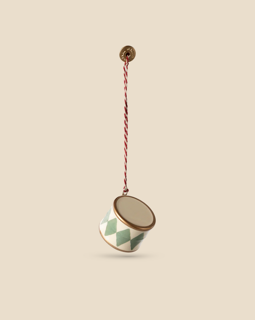 Against a beige background, the Maileg Metal Ornament, Small Drum in Mint hangs by a twisted red and white cord, showcasing its charm with hand-painted details in a striped green and white pattern.