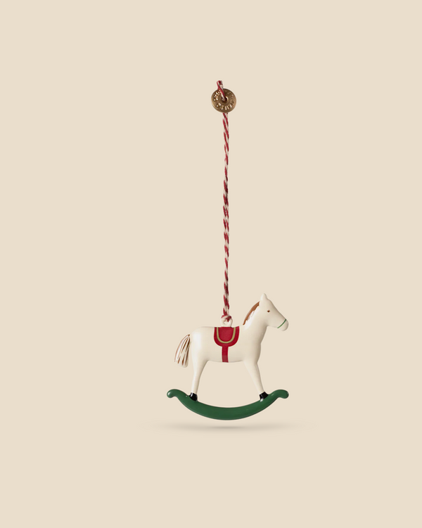 The Maileg Metal Ornament, Rocking Horse with a red saddle and striped hanging string sways gently against a beige background. This charming piece, featuring handpainted details and a simple design, rests on a green base.