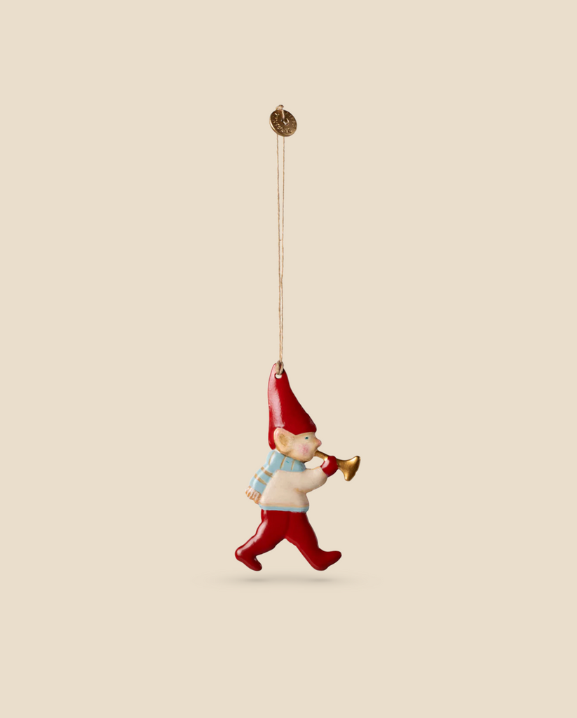 The Maileg Metal Ornament, Pixy Trumpet, is a delightful gnome figurine set against a beige background. Featuring handpainted details, the gnome wears a cheerful red hat and boots, joyfully marching while holding a small horn.