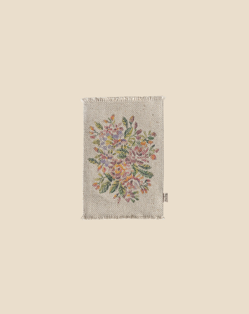 The Maileg Flower Rug, Medium, presents a delightful flower pattern with pink, yellow, and orange blossoms nestled among green leaves on a beige backdrop. Ideal for dollhouses, it is adorned with fringed edges and includes a simple tag on the side. (Ships in approximately one week.)