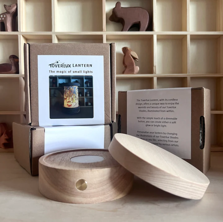 Wooden shelves feature animal figurines and multiple packages of the Toverlux Lantern, one of which showcases an image of the cordless, dimmable lantern. The wooden base with a cylindrical cap is prominently displayed next to a partially visible description card.