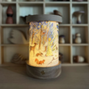 The Toverlux Lantern, with its customizable shade showcasing a woodland scene of an angel, deer, squirrel, and trees, casts a gentle glow throughout the room. The backdrop accentuates a wooden shelf decorated with organic figures and forms. (Please note that shipping takes approximately one week.)