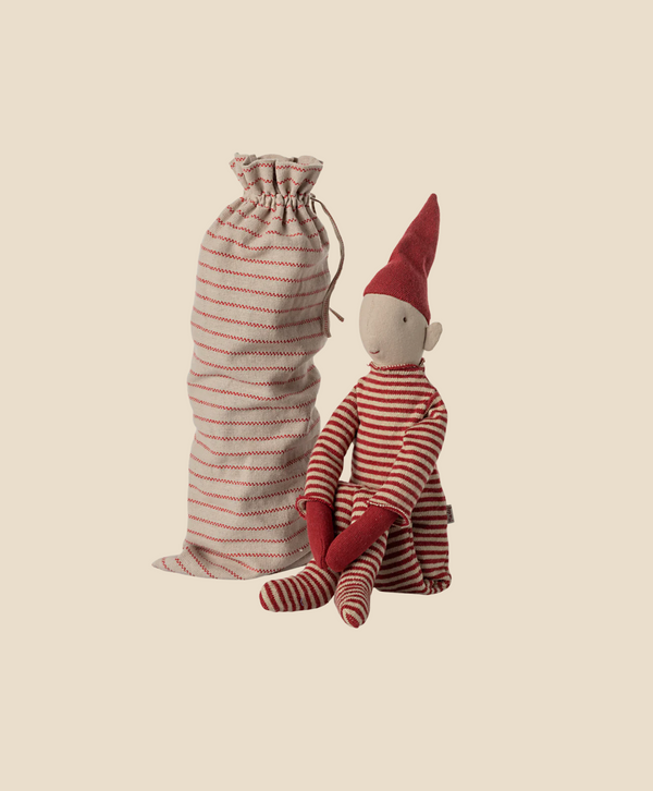 The Maileg Long Legged Pixy, Large, dressed in a whimsical red and white striped outfit with a pointed red hat, is accompanied by a matching drawstring bag. A warm beige background enhances the charm of the scene.