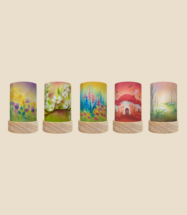 A set of five decorative Toverlux Lantern Shades, known as the Seasons collection by Baukje Exler, includes nature-themed designs such as sunflowers, leaves, lupines, mushroom houses, and a forest path. Each lantern in this set features Magic Silhouettes on a wooden base in a cylindrical shape against a beige background. The set ships in approximately one week.