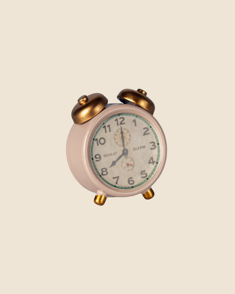 A small, vintage-style alarm clock sits beside the Maileg Micro Bunny Bedroom Set, featuring charming bunny and accessories, all set against a plain beige background with the clock showing approximately 12:00.