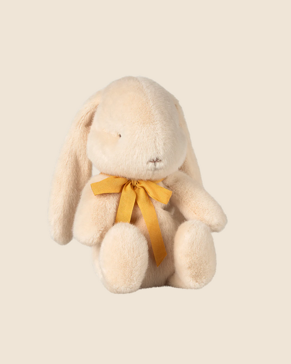 The small cream Maileg Plush Bunny, available for pre-order and shipping in February, is crafted from soft plush fabric and recycled polyester. It has a sweet expression, floppy ears, and a yellow ribbon around its neck against a plain beige background.